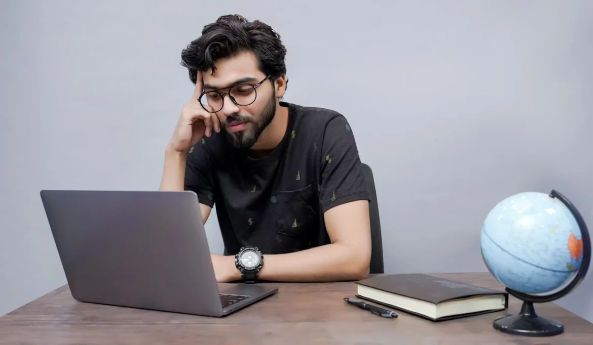 A man in glasses is at a desk, engaged with his laptop