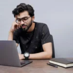 A man in glasses is at a desk, engaged with his laptop
