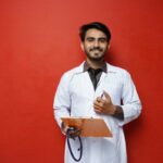 A male doctor in a white coat holding a clipboard and a stethoscope