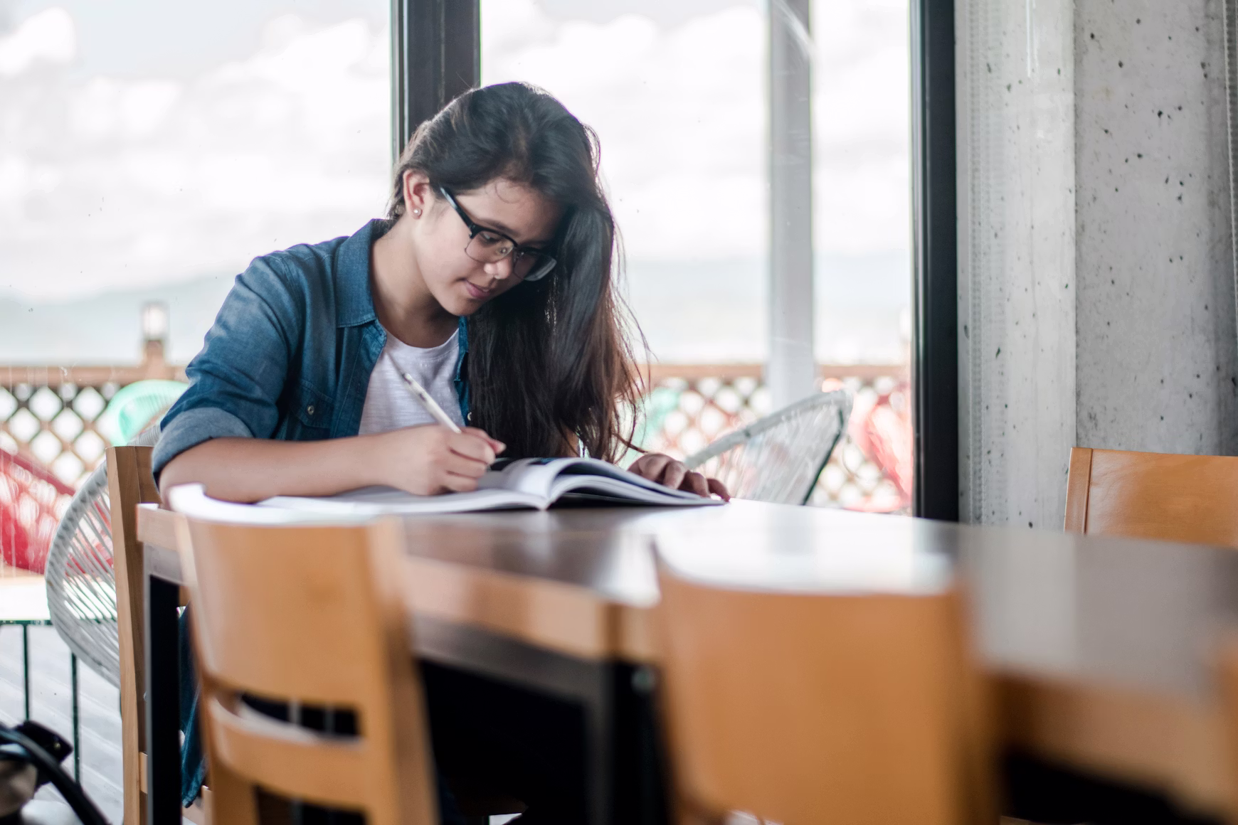 A bright, mid-shot photograph captures a young woman sitting at a smooth, dark wooden table, focused on writing in an open notebook or textbook.