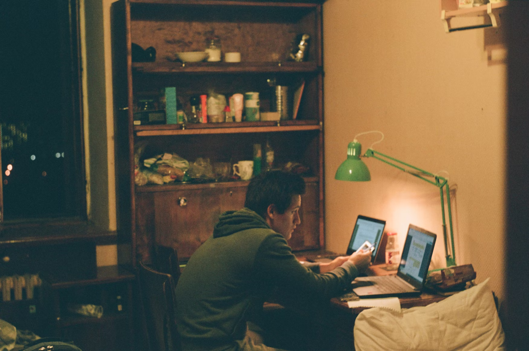 A candid, low-light photograph captures a young person seated at a wooden desk in an apartment at night.