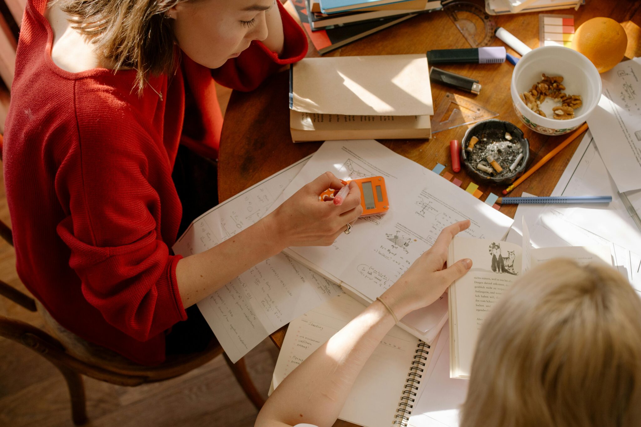 An overhead shot of two people sitting at a wooden table covered in papers, books, and study materials.