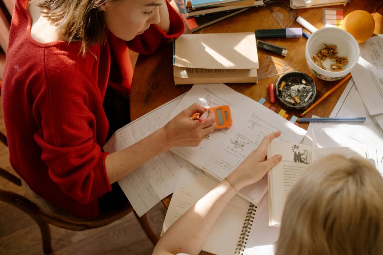 An overhead shot of two people sitting at a wooden table covered in papers, books, and study materials.