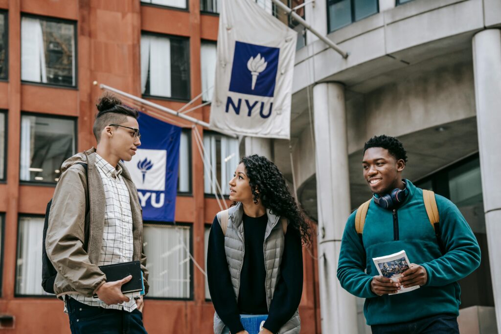 Three young college students are standing and talking outside a brick university building. Two flags with the NYU logo are visible behind them.