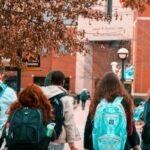 A group of college students with backpacks walking away from the camera toward a brick university building on a fall day. 